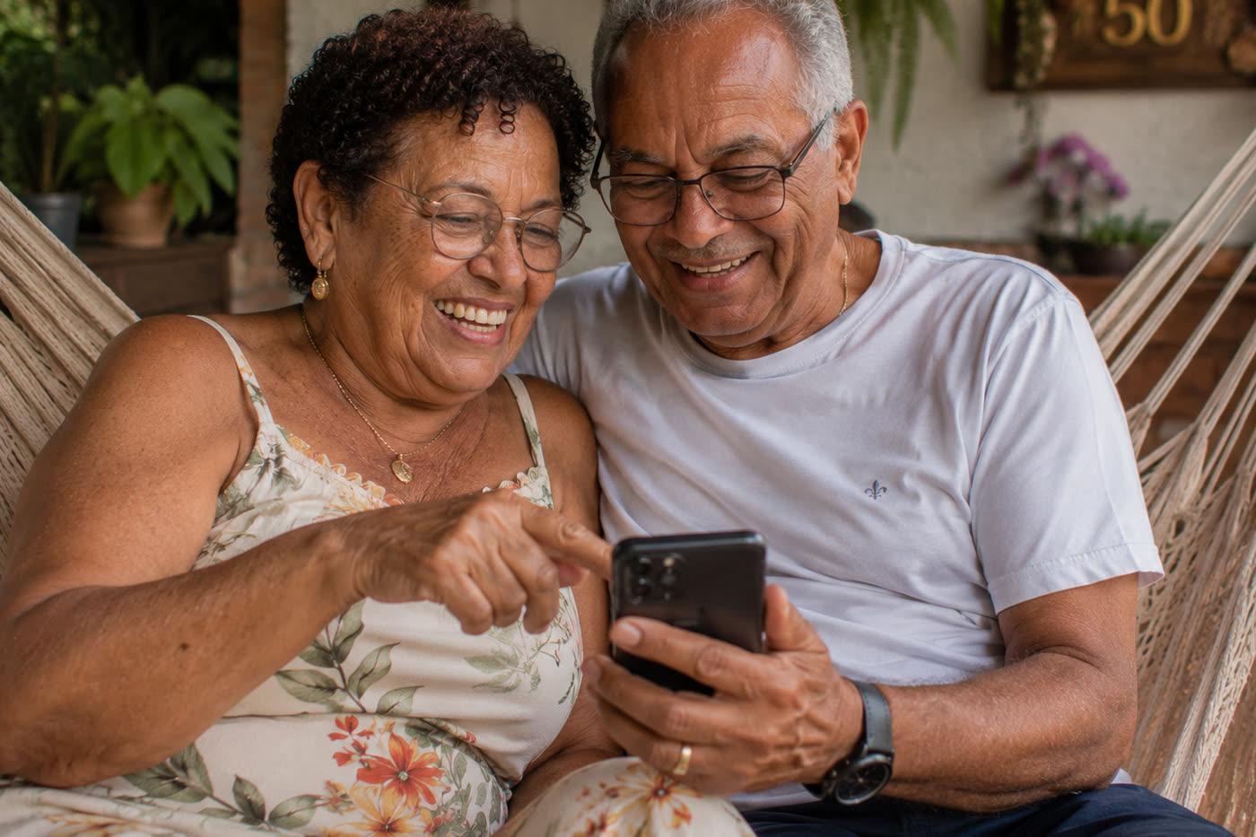 Casal de bodas sentado na rede da varanda, apontando para o celular e rindo juntos