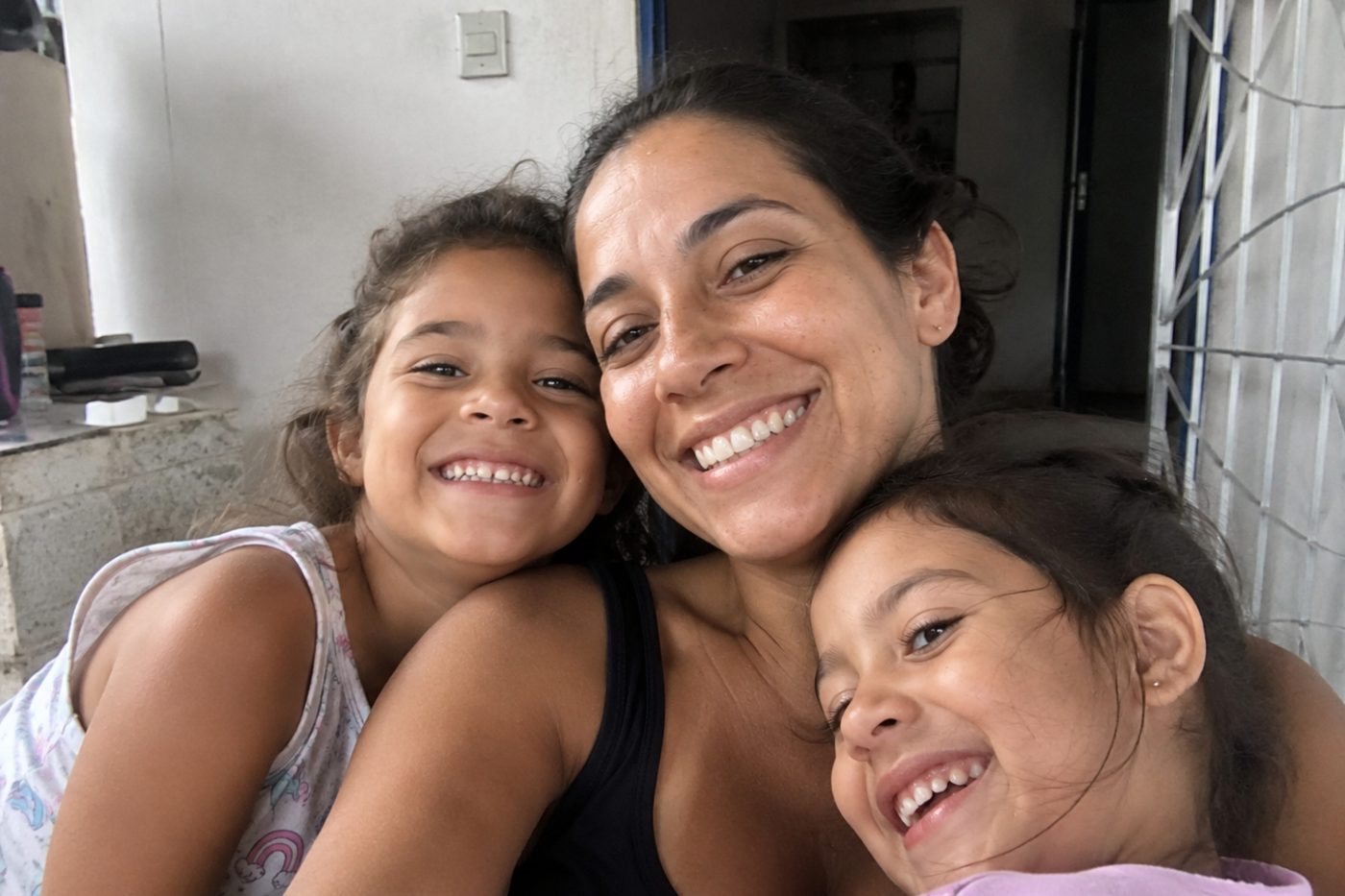 Mãe abraçada com as duas filhas, as três rindo juntas em casa