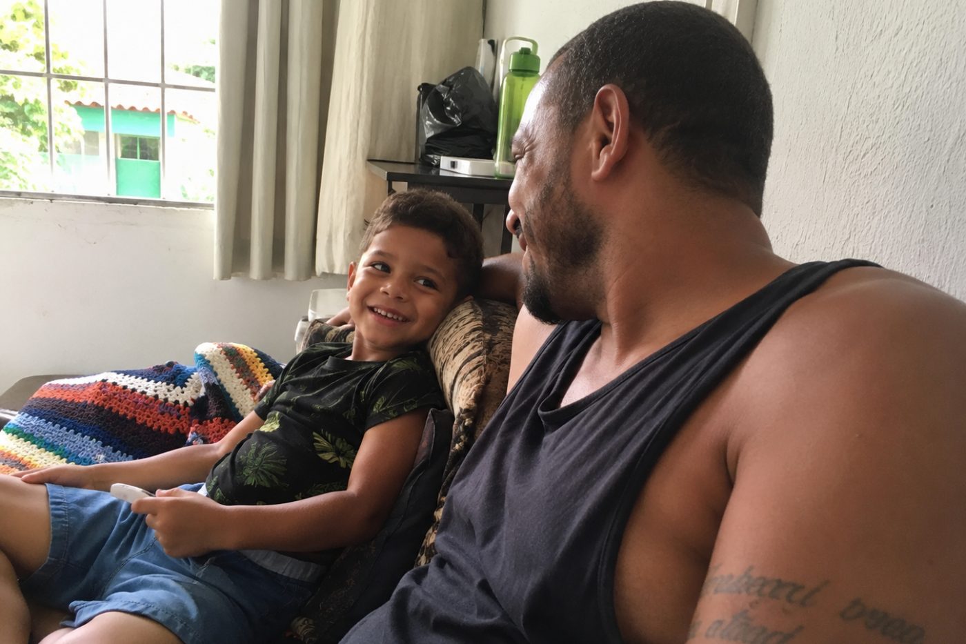 Pai e filho pequeno no sofá de casa, rindo um para o outro