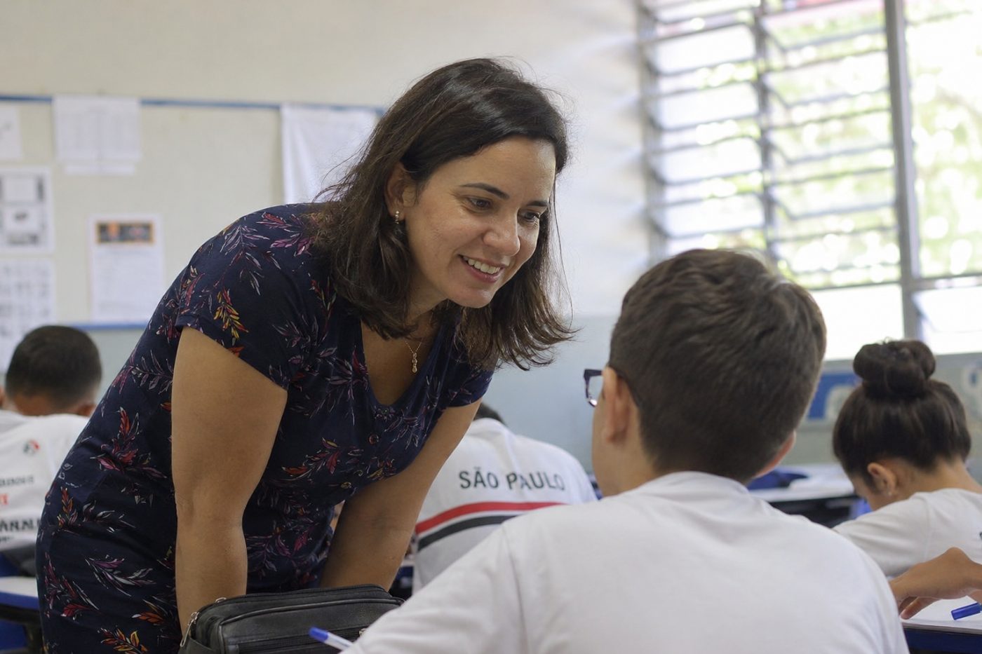Professora inclinada sobre a carteira, conversando e sorrindo com um aluno na sala de aula