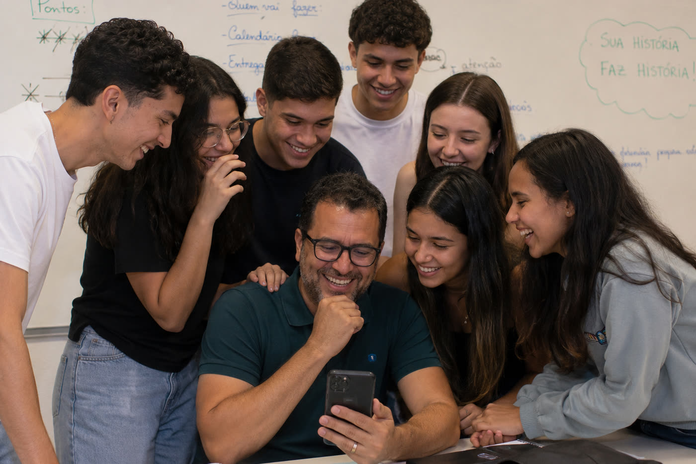 Professor cercado pelos alunos na sala, todos rindo do que ouvem no celular dele